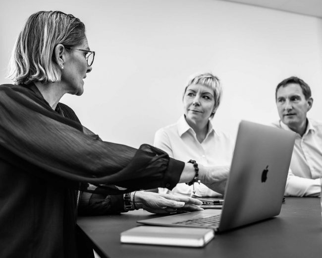 two women and a man around a table with a macbook open